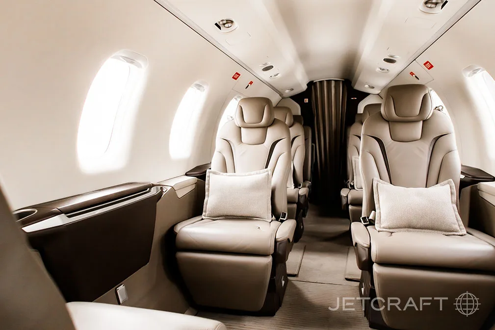 The aircraft cabin features light brown leather seats with small pillows, arranged in a club configuration.