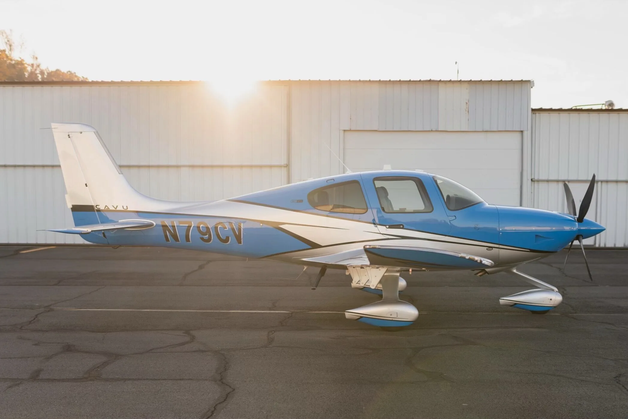 A blue and silver Cirrus SR22T GTS aircraft, registration N79CV, parked on an airfield with hangars in the background and sun glare.