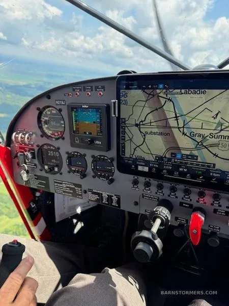 A cockpit view showing the instrument panel with a tablet displaying a map and an outside view of the ground from an aircraft in flight.