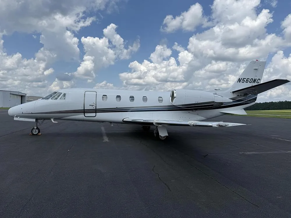 A white Cessna Citation private jet with black and grey stripes parked on an asphalt tarmac under a cloudy sky.