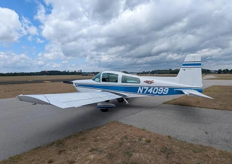 A white and blue Grumman AA-5 Traveler aircraft with registration N74099 parked on an asphalt runway under a cloudy sky.