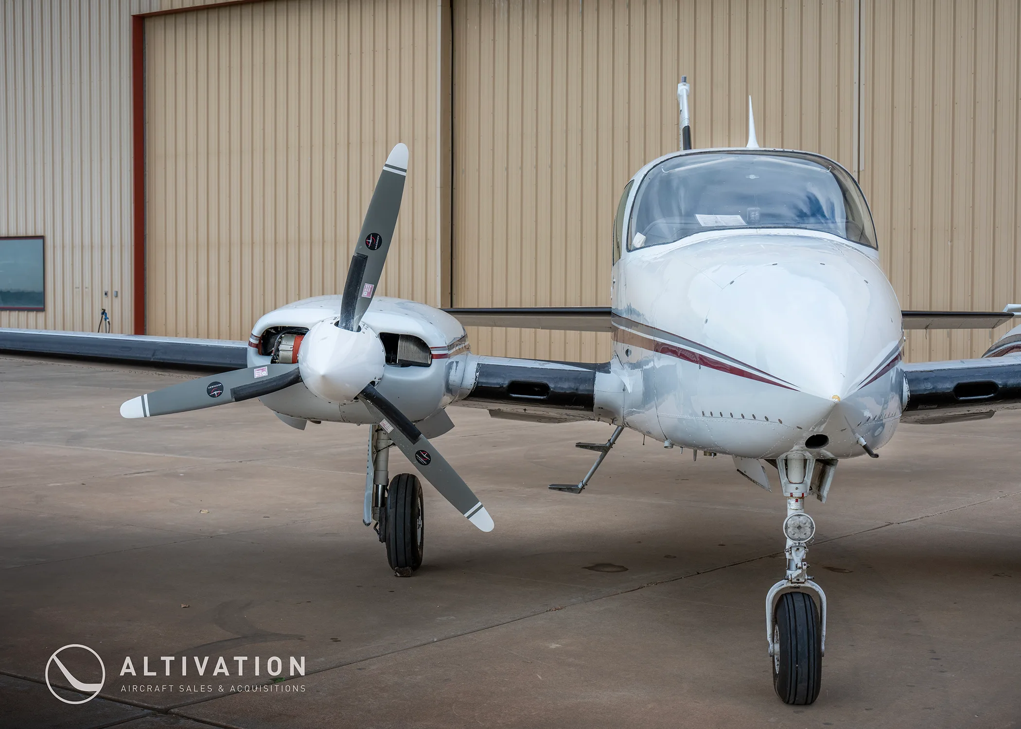 A front view of a white twin-engine propeller aircraft with red and grey stripes, parked on an airfield.