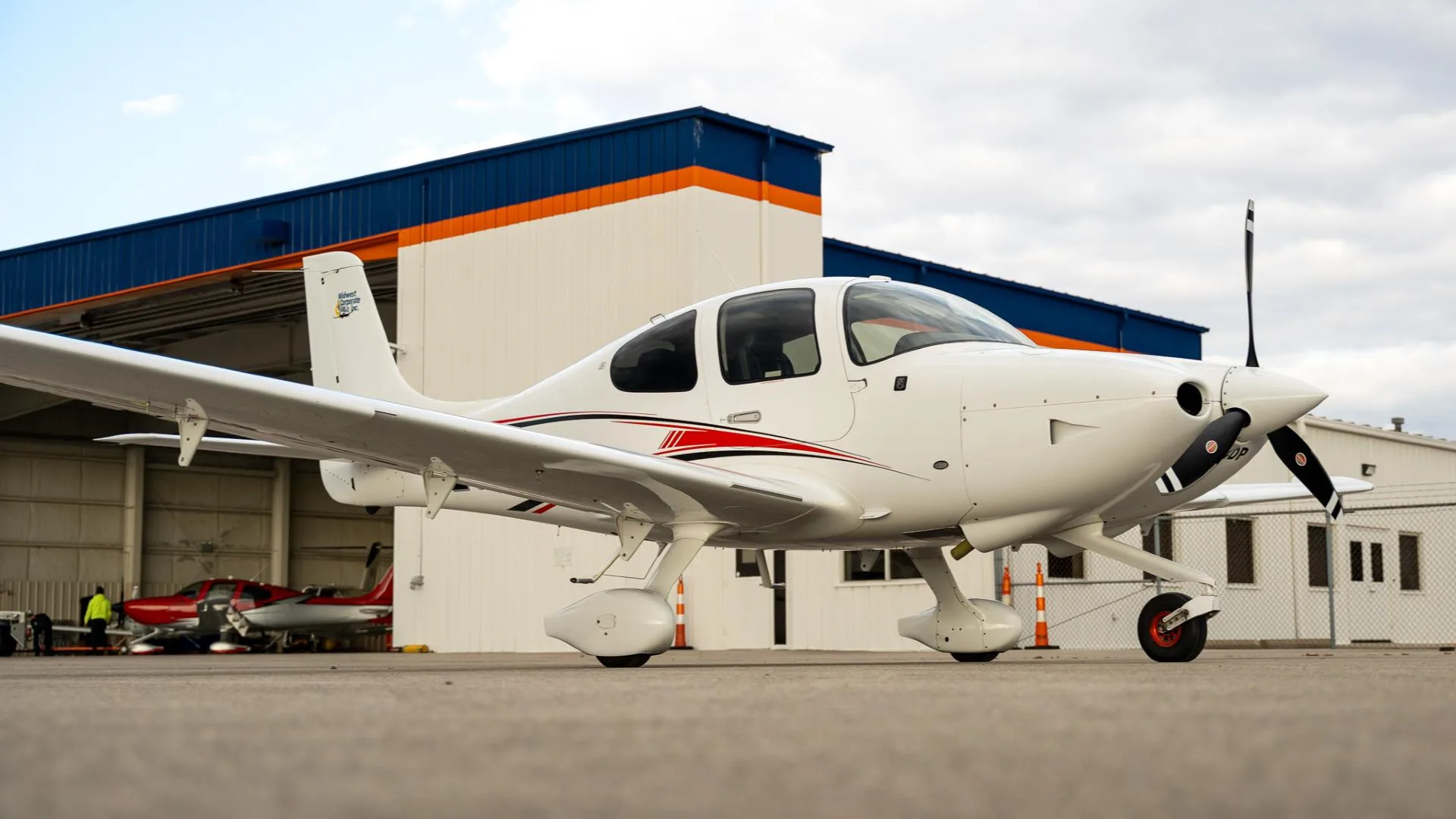 A white Cirrus SR22 aircraft with red and black stripes parked on a tarmac in front of a hangar.