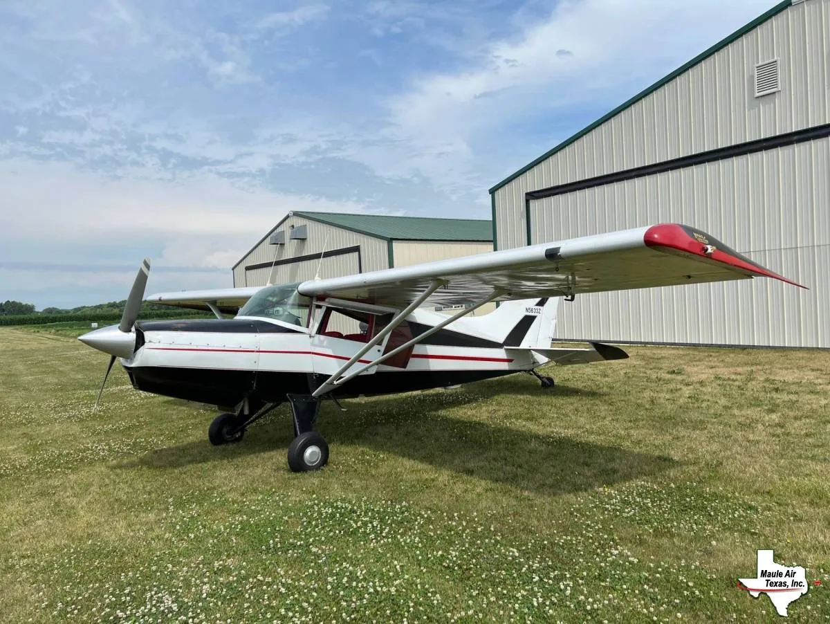 A white and black Maule M-5-235C Lunar Rocket with red wingtips is parked on a grassy field in front of hangars.