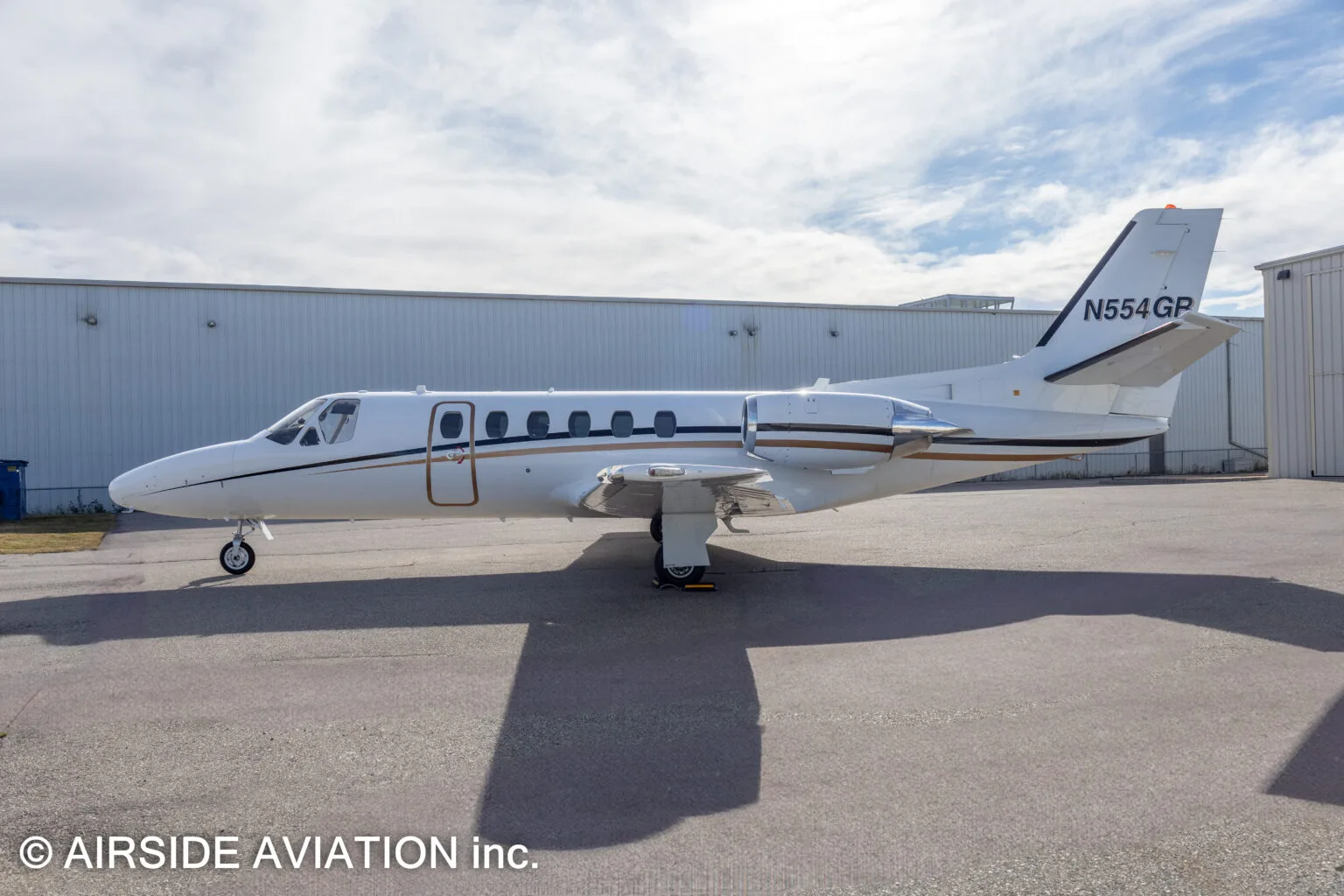 A white Cessna Citation business jet with gold and black stripes, registration N554GR, parked on an airfield next to hangars.