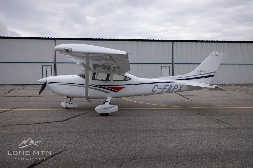 A white Cessna 182 Skylane with grey and blue stripes, registration C-FAPX, parked on an asphalt ramp in front of hangars.