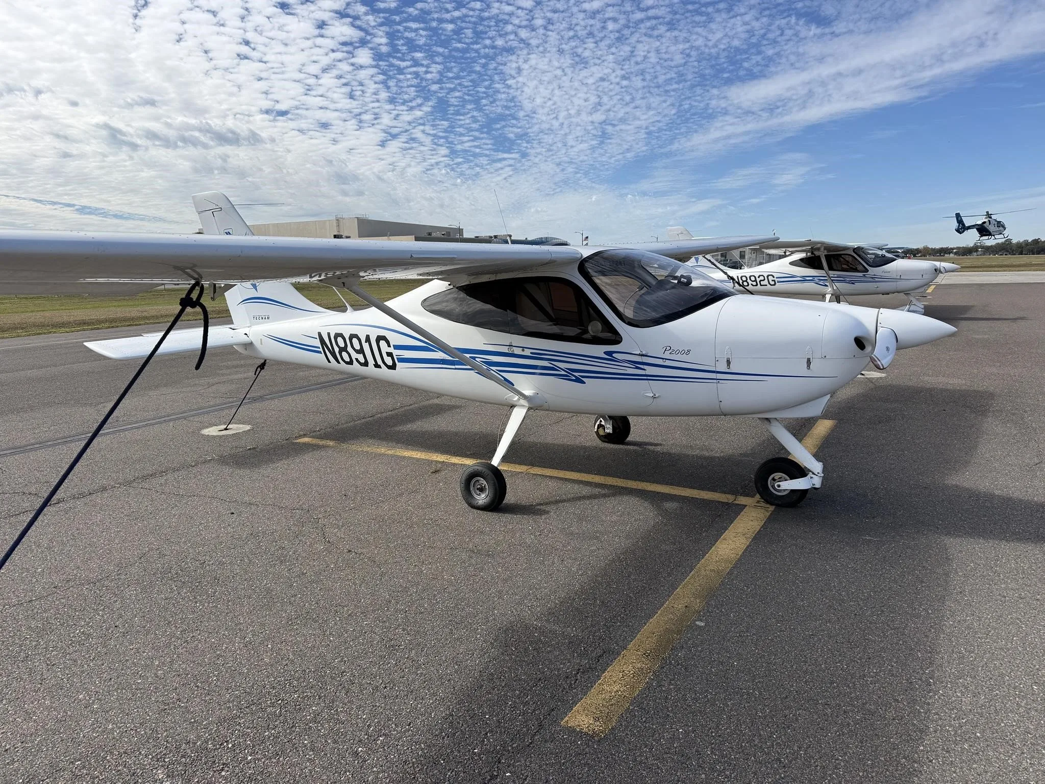 A white Tecnam P2008 aircraft with blue stripes, registration N891G, parked on an airport tarmac under a partly cloudy sky.