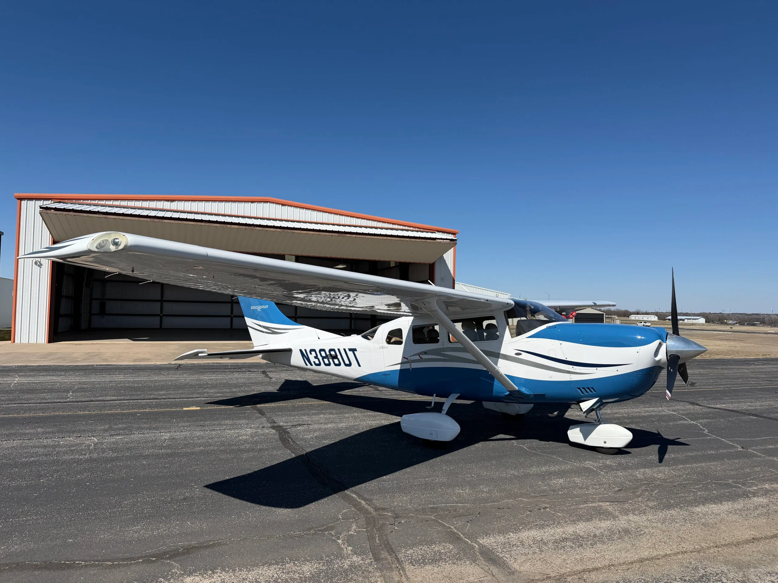 A white and blue single-engine propeller aircraft with registration N388UT is parked on an asphalt ramp in front of an open hangar under a clear blue sky.