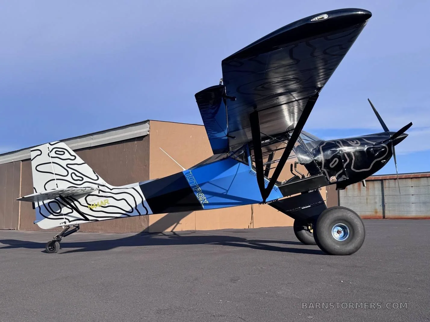 A black, blue, and white Kitfox aircraft with abstract patterns on the tail and nose, parked on an asphalt tarmac.