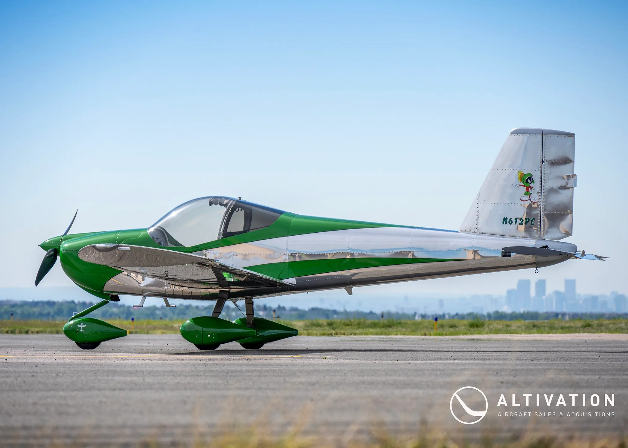 A side view of a green and polished aluminum Vans RV-12 aircraft, registration N612PC, parked on an airfield with a city skyline in the distance.