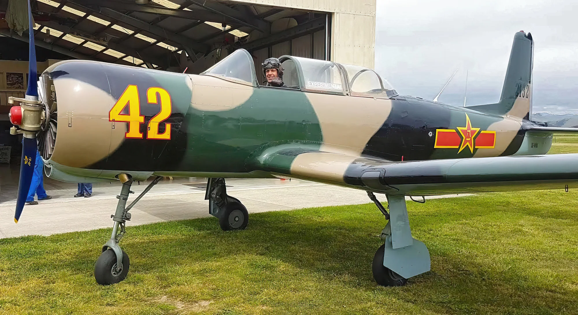 A single-engine Nanchang CJ-6 aircraft with a camouflage paint scheme, yellow "42" on the fuselage, and a pilot in the cockpit, parked on a grassy airfield.