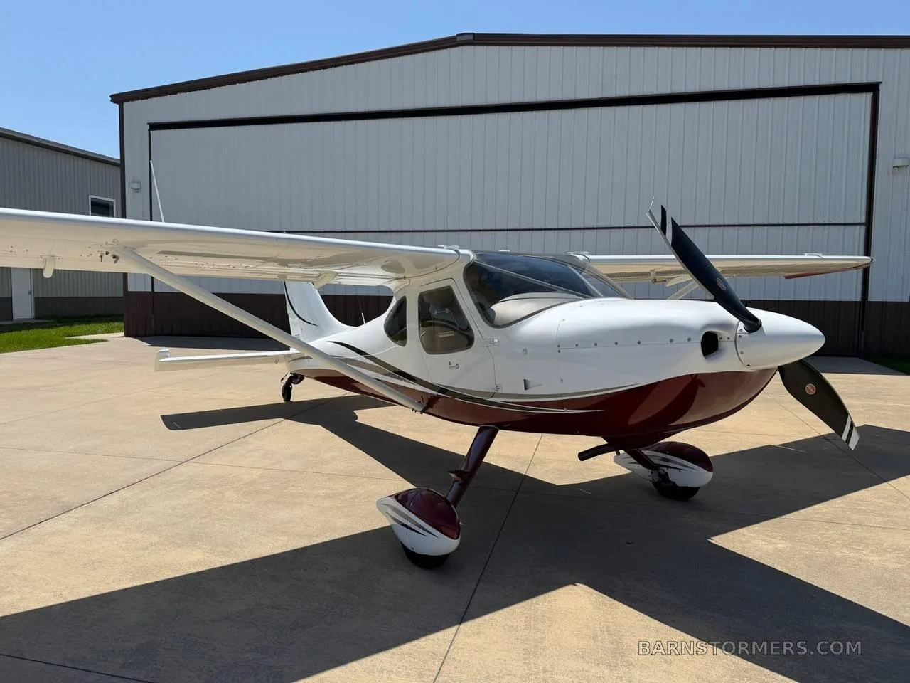 A white and maroon high-wing single-engine aircraft is parked on a concrete tarmac in front of hangars.