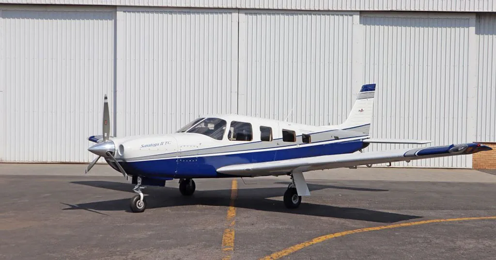 A white and blue Piper Saratoga II TC aircraft parked on an asphalt ramp with a hangar in the background.