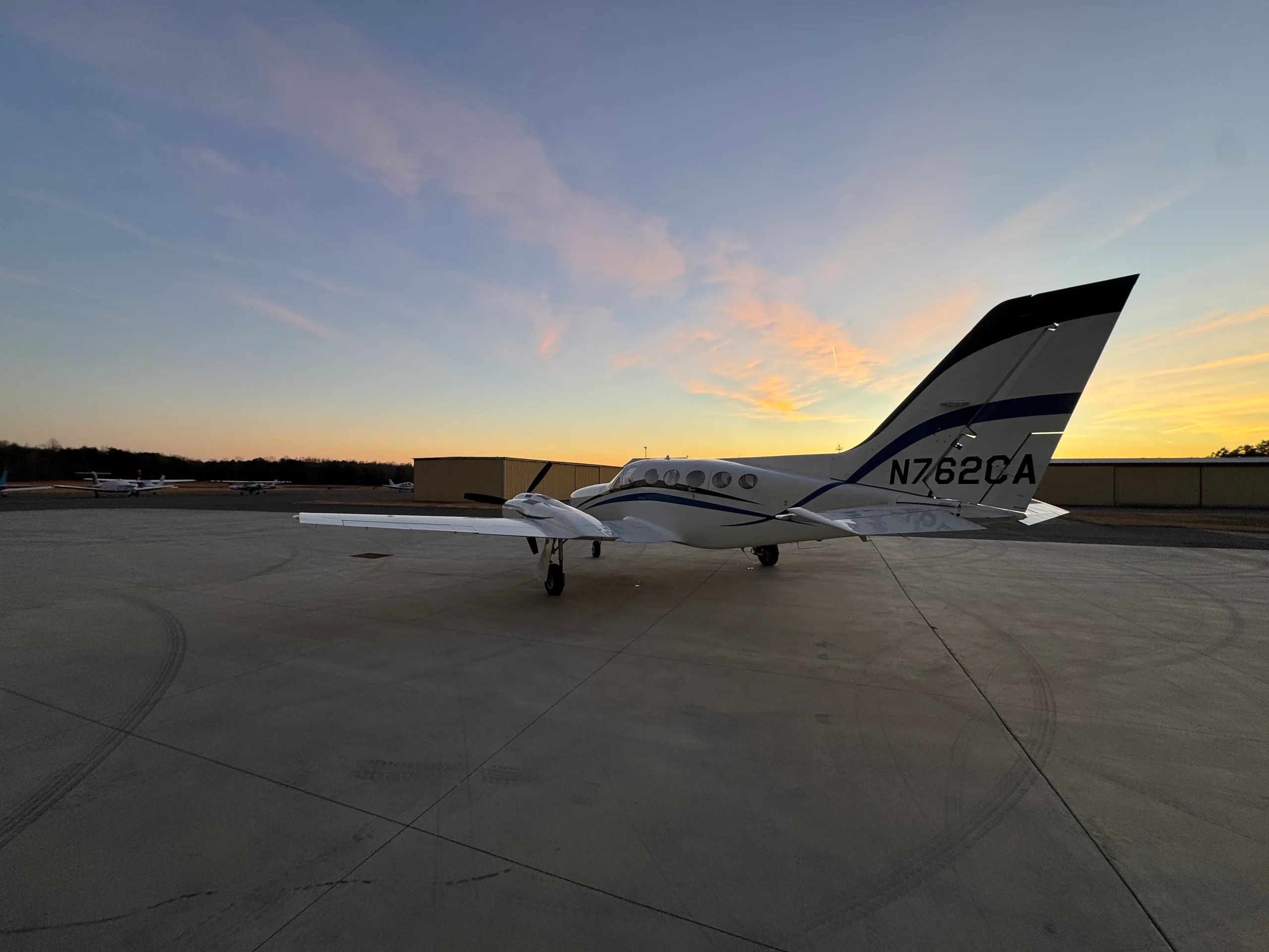 A white twin-engine aircraft with blue stripes, N762CA, parked on an airfield at sunset.
