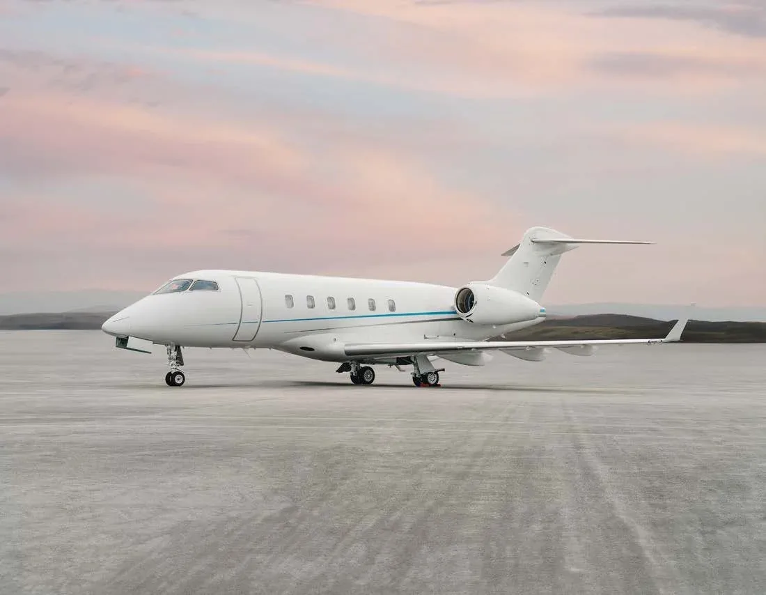 A white business jet with a blue stripe is parked on an airfield under a pastel-colored sky.
