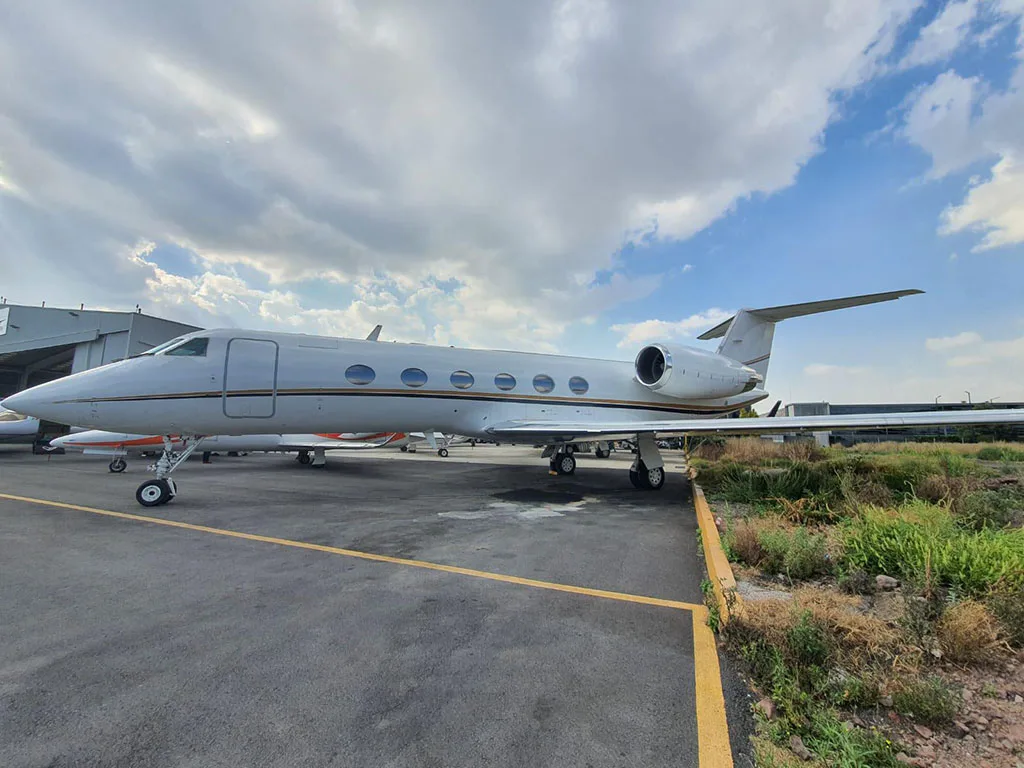 A white business jet with gold and black stripes parked on an asphalt apron under a cloudy sky.
