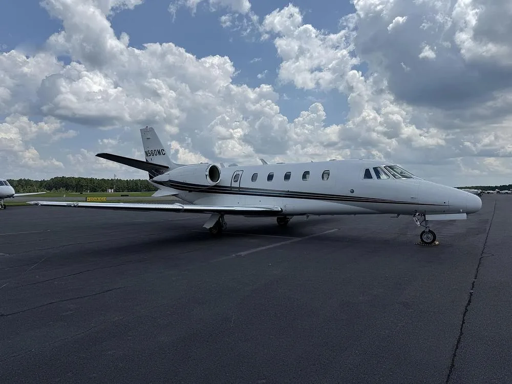 A side view of a white Cessna Citation private jet with black and grey stripes parked on an airport tarmac under a cloudy sky.