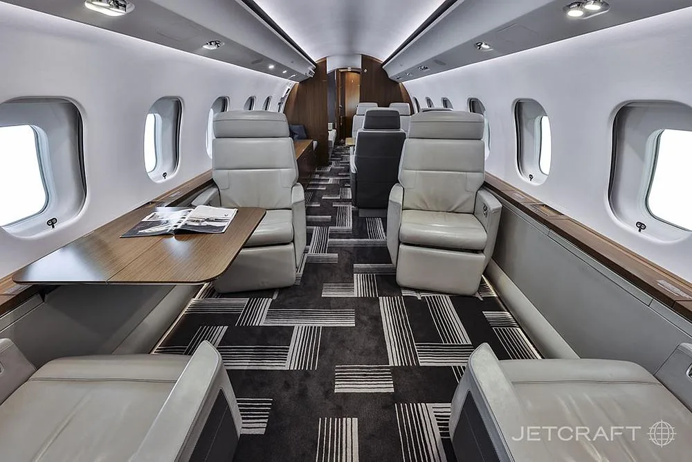 A luxurious aircraft cabin interior with light grey leather seats, a wooden table, and a dark patterned carpet.