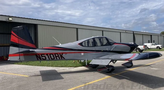 A gray and red single-engine propeller aircraft, N510RK, is parked on an airfield in front of a hangar.