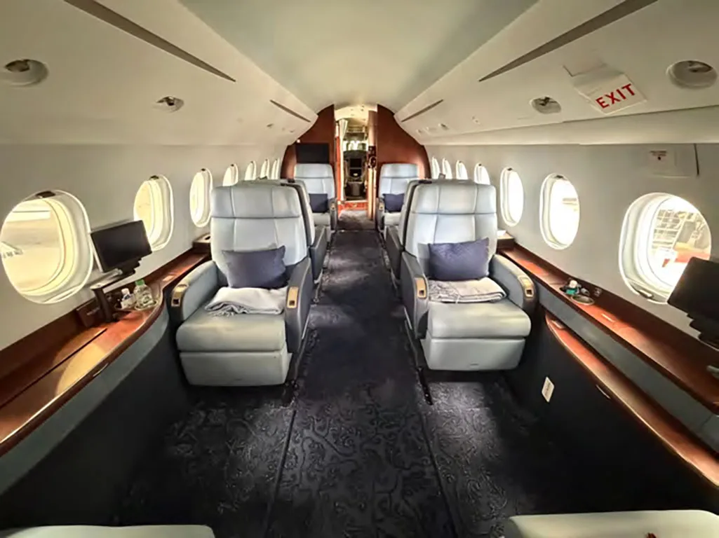The luxurious cabin interior of a business jet, featuring light blue leather seats, dark blue patterned carpet, and wooden accents.