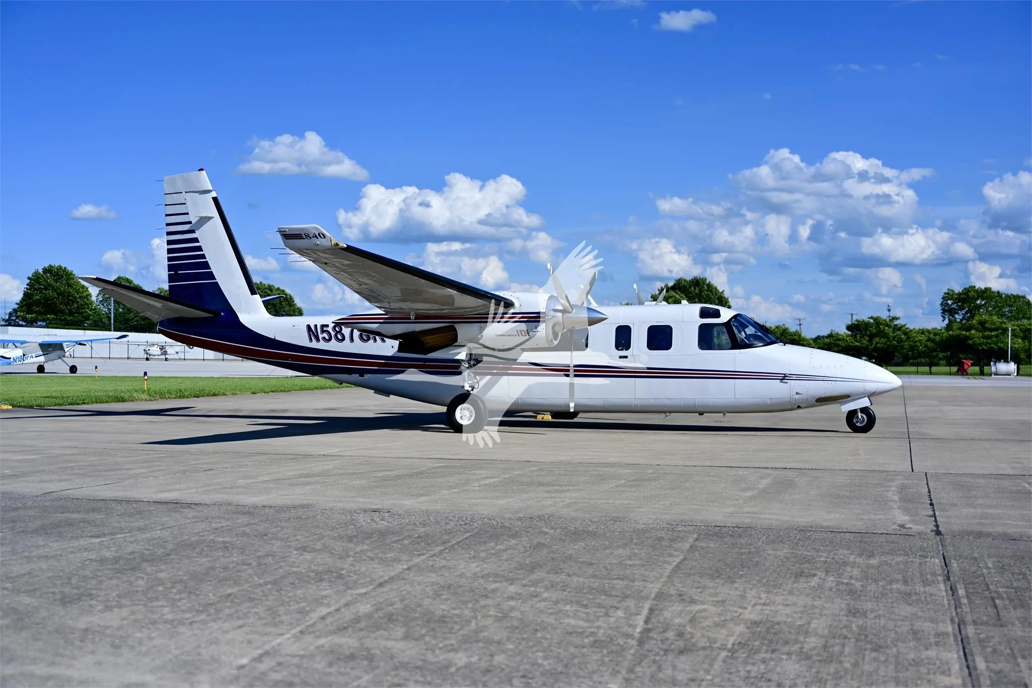A white and blue Commander 840 twin-engine turboprop aircraft is parked on a tarmac under a blue sky.