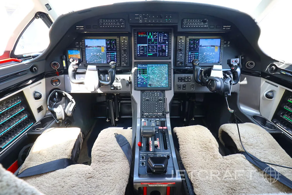 The modern glass cockpit of a Pilatus PC-12 aircraft with multiple display screens and sheepskin seat covers.