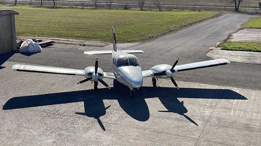 A silver twin-engine propeller aircraft with red stripes on the tail and wings is parked on an asphalt apron, casting long shadows.