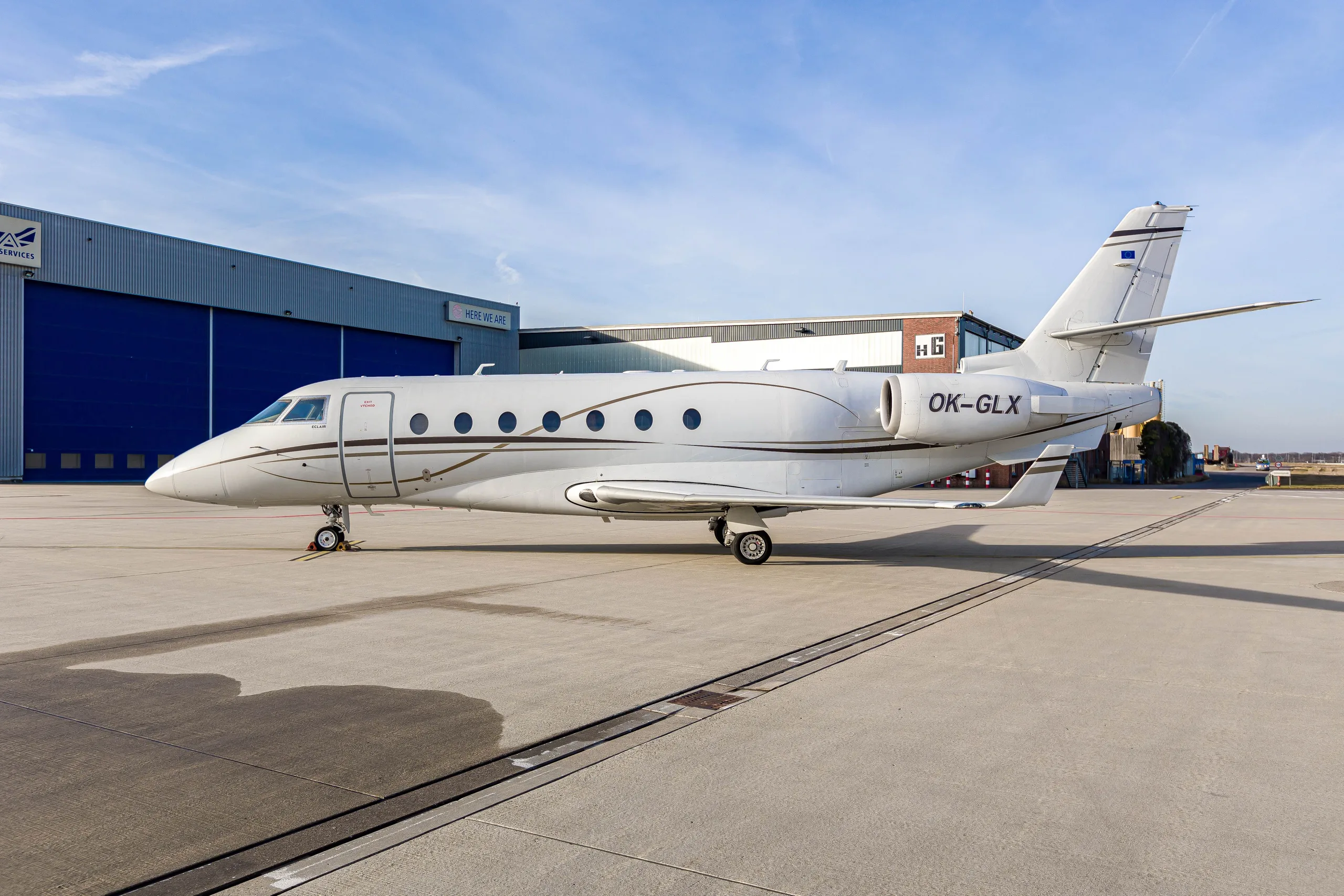 A white Gulfstream G200 business jet with registration OK-GLX parked on an airport tarmac next to a blue hangar.