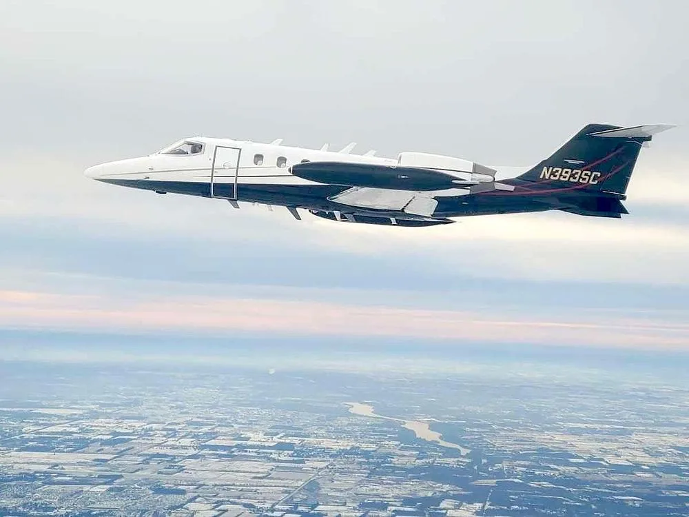 A Learjet 35A with a white and black paint scheme flying over a landscape with fields and a river.