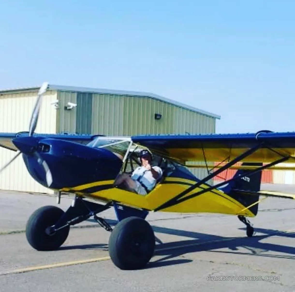 A blue and yellow high-wing aircraft with large tundra tires and a pilot in the cockpit, on an airfield with a hangar in the background.