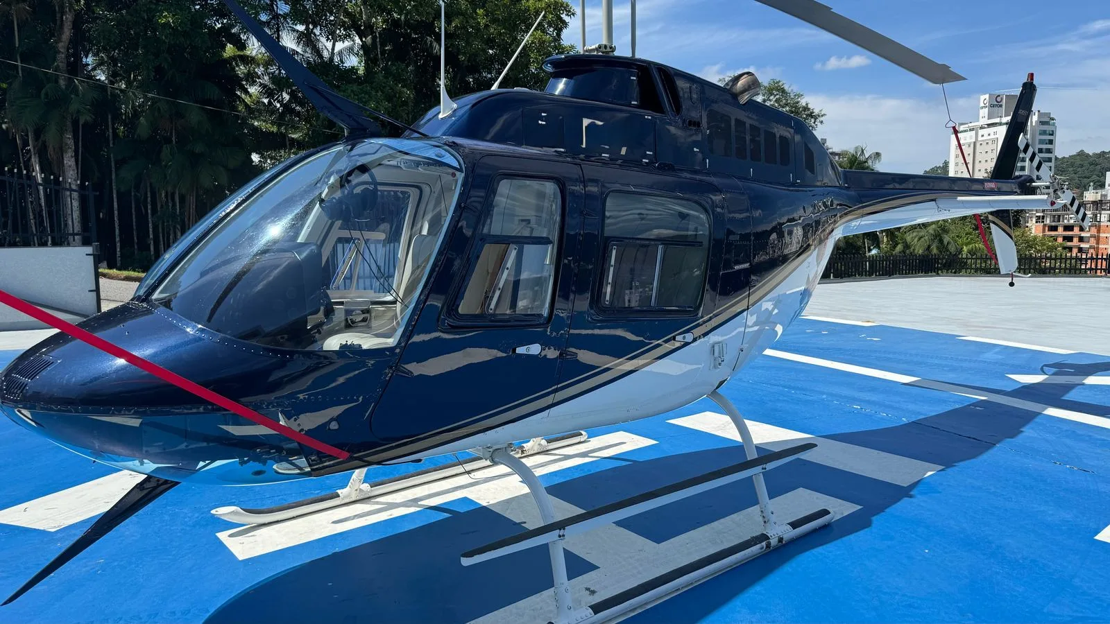 A dark blue and white helicopter with a red tie-down strap on a blue helipad under a clear sky.