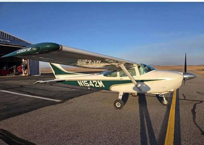 A white and green Cessna aircraft (N1542M) is parked on an asphalt runway next to a hangar under a clear blue sky.
