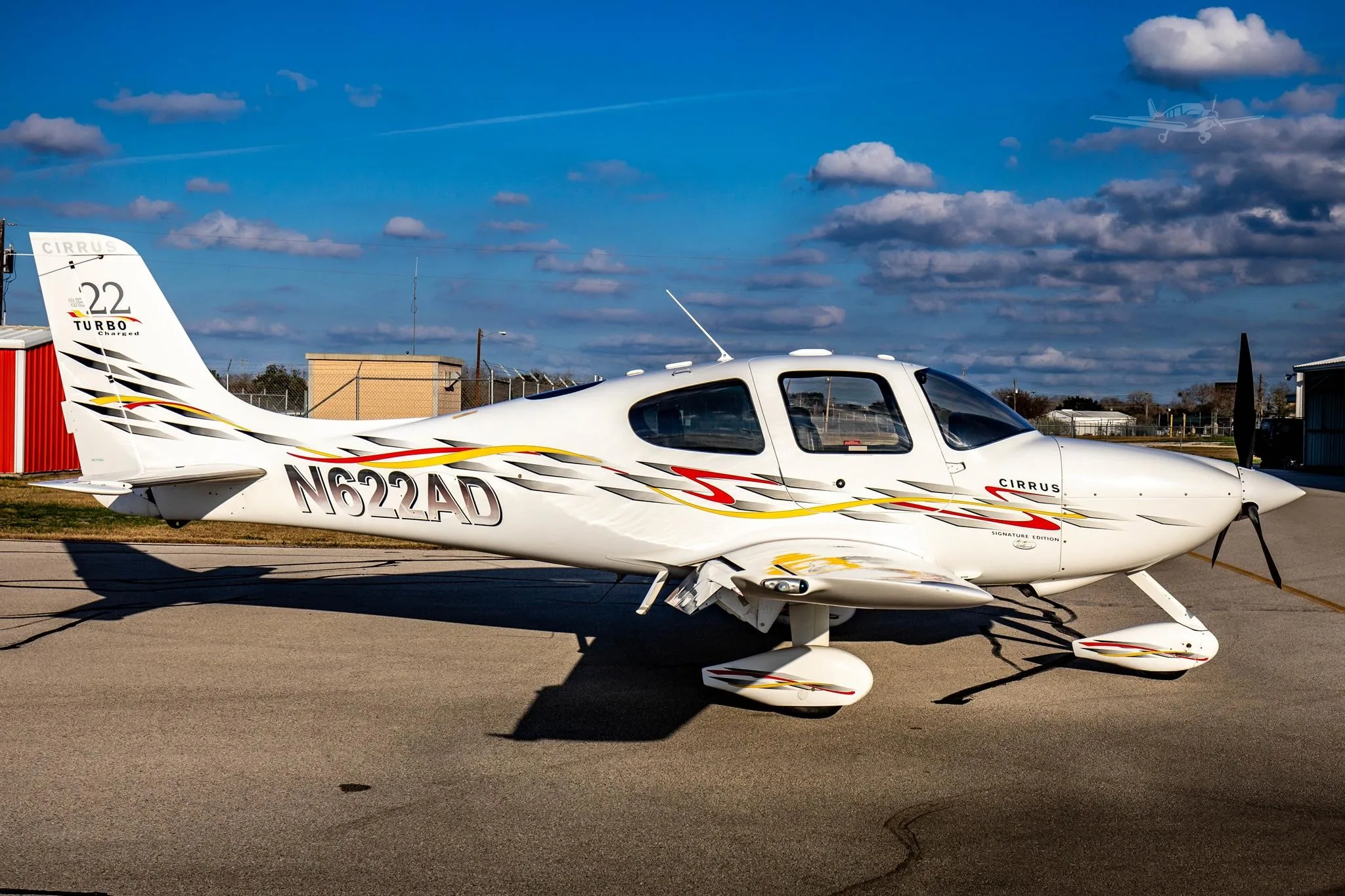 A white Cirrus SR22 Turbo Charged aircraft with registration N622AD, featuring red, yellow, and black stripe livery, parked on an asphalt ramp under a blue sky with clouds.