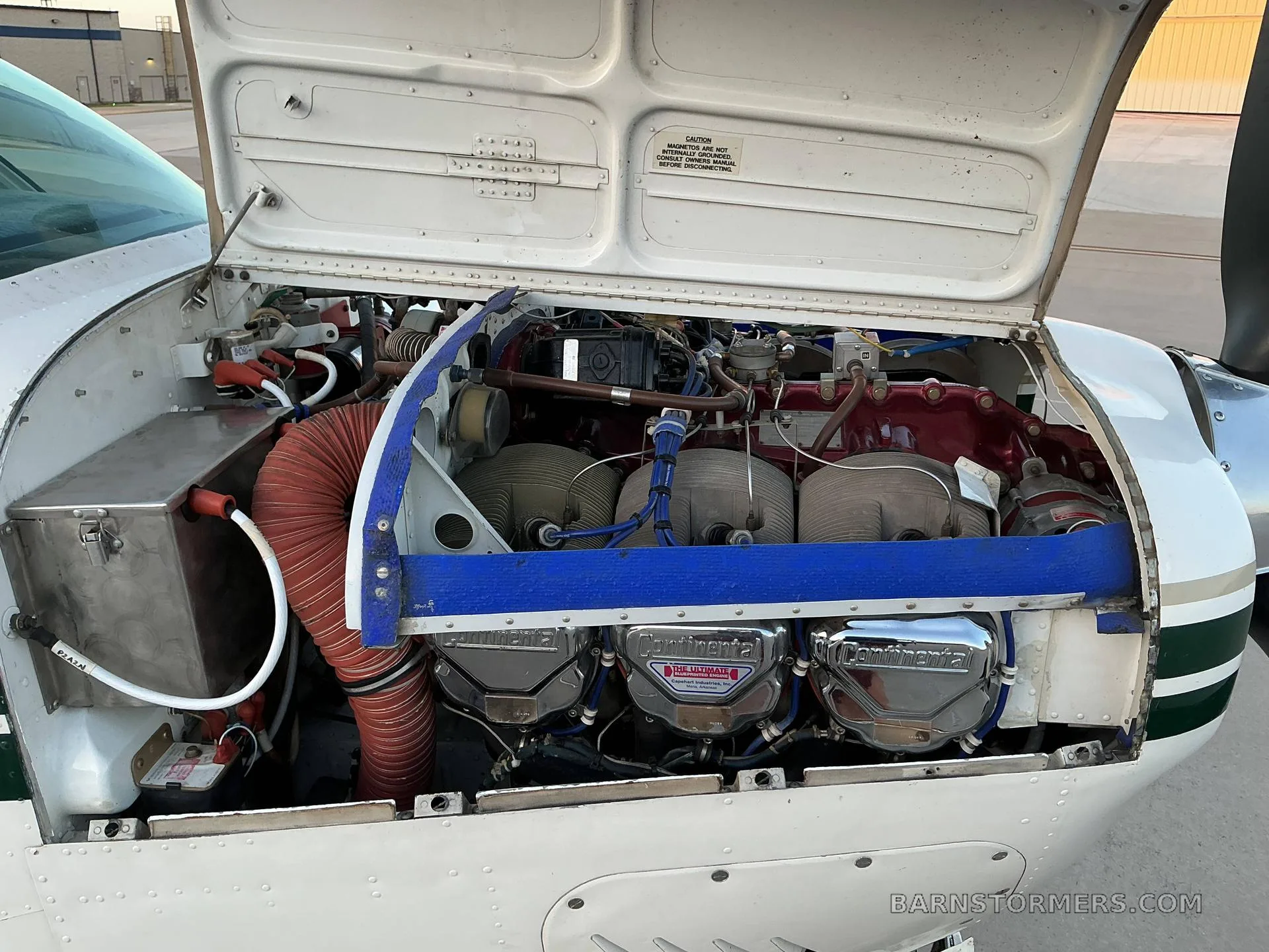 The open engine cowling of a white aircraft, revealing a Continental engine with red and blue components.