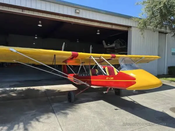 A red and yellow ultralight aircraft, possibly an Air-Cam, is parked outside a hangar on a sunny day.