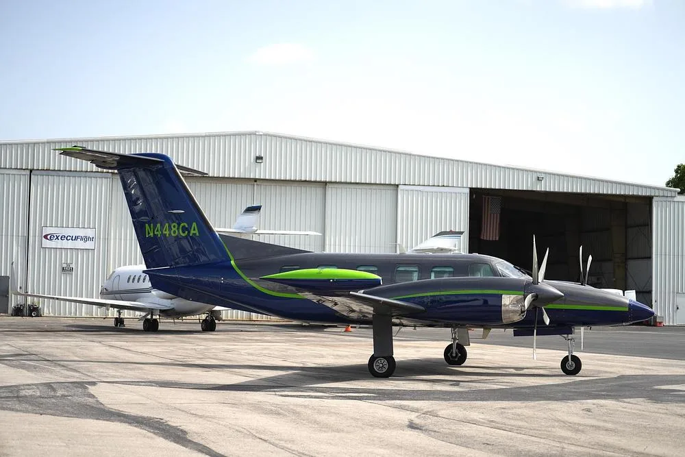 A side view of a dark blue and grey twin-engine turboprop aircraft with a green stripe, tail number N448CA, parked in front of a hangar.