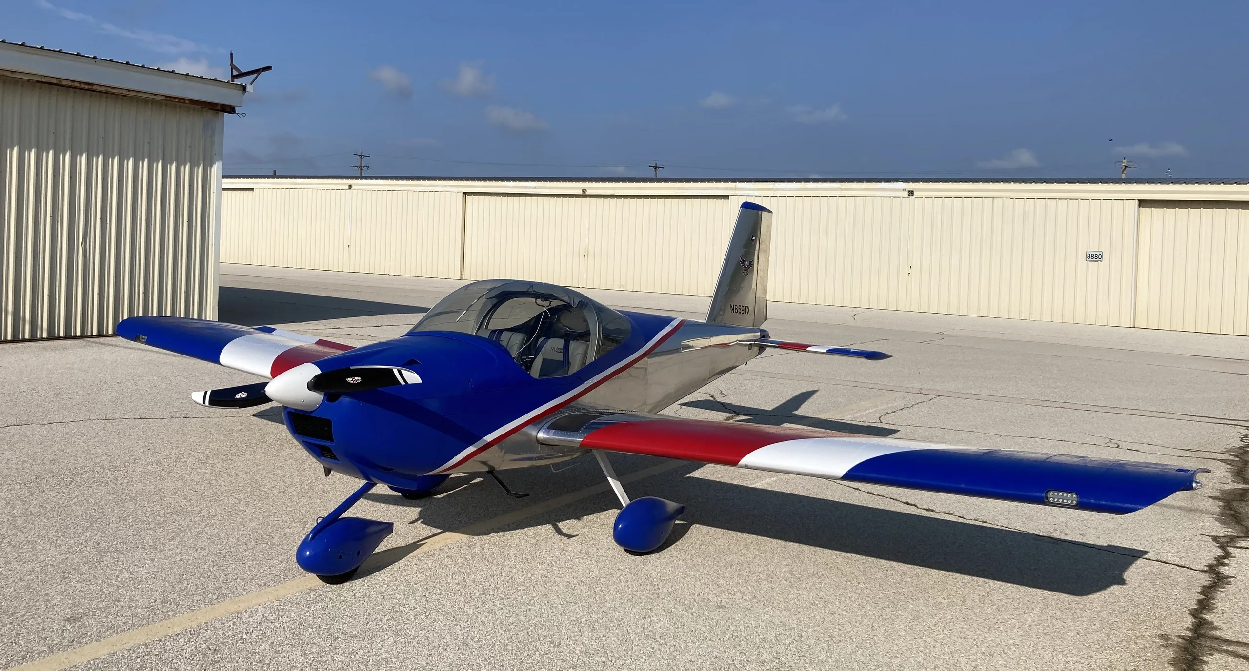 A blue, silver, red, and white Van's RV-7 aircraft is parked on an airfield ramp next to hangars under a clear sky.