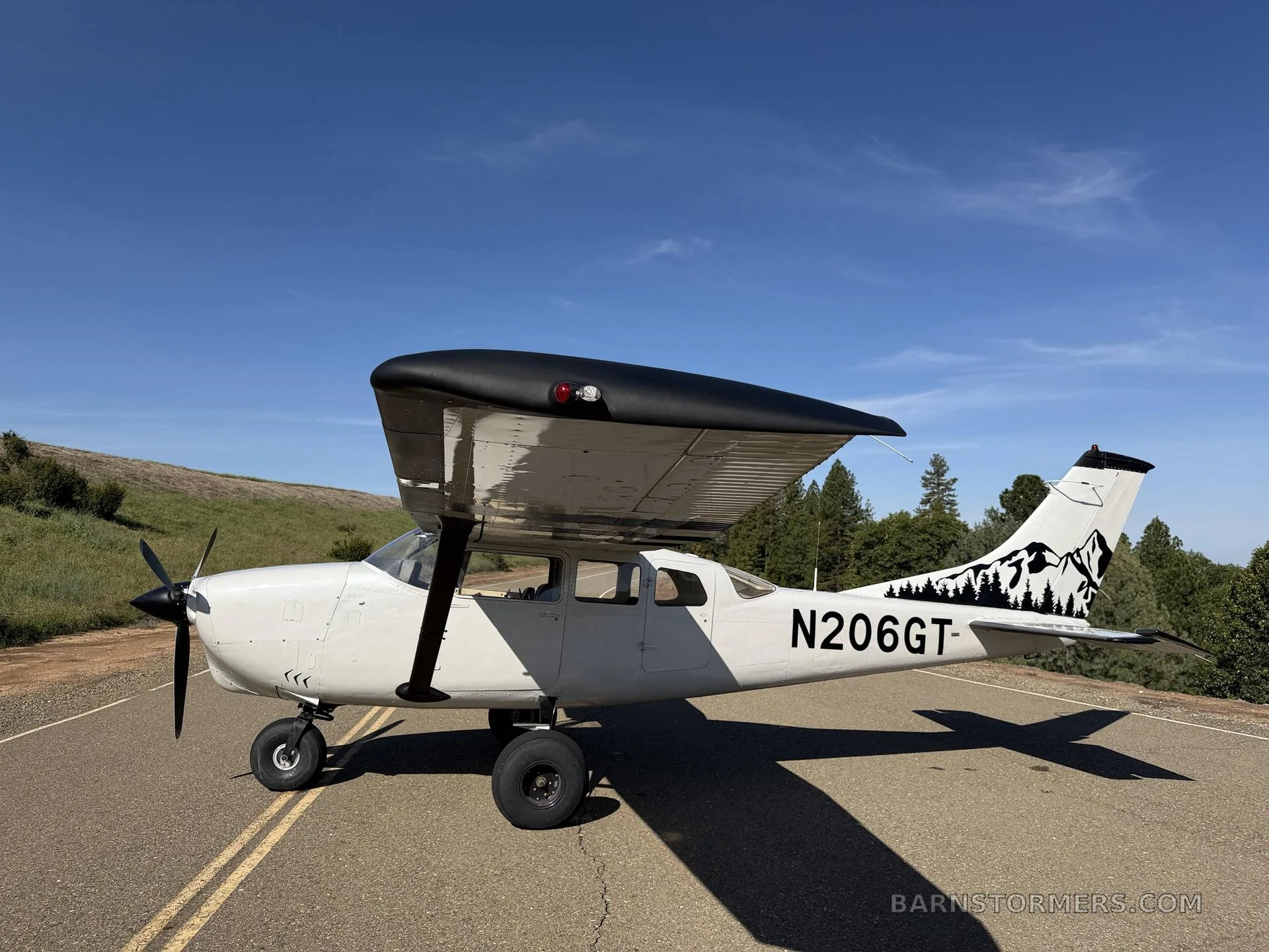 A white Cessna 206GT with black wings and mountain tail art, parked on a paved surface under a clear blue sky.