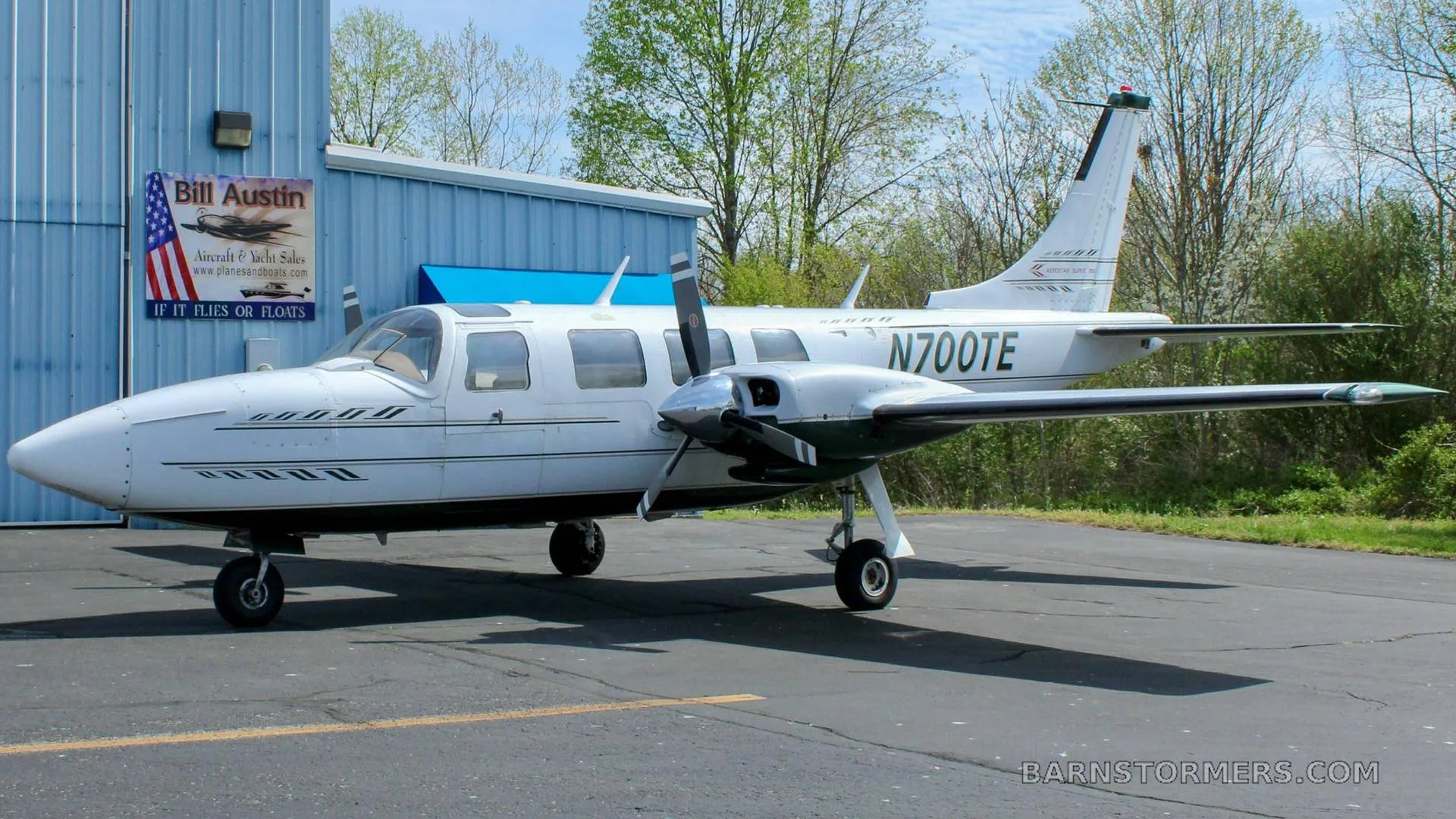 A white Piper Aerostar twin-engine aircraft with black stripes, registration N700TE, parked on an asphalt ramp next to a blue hangar.