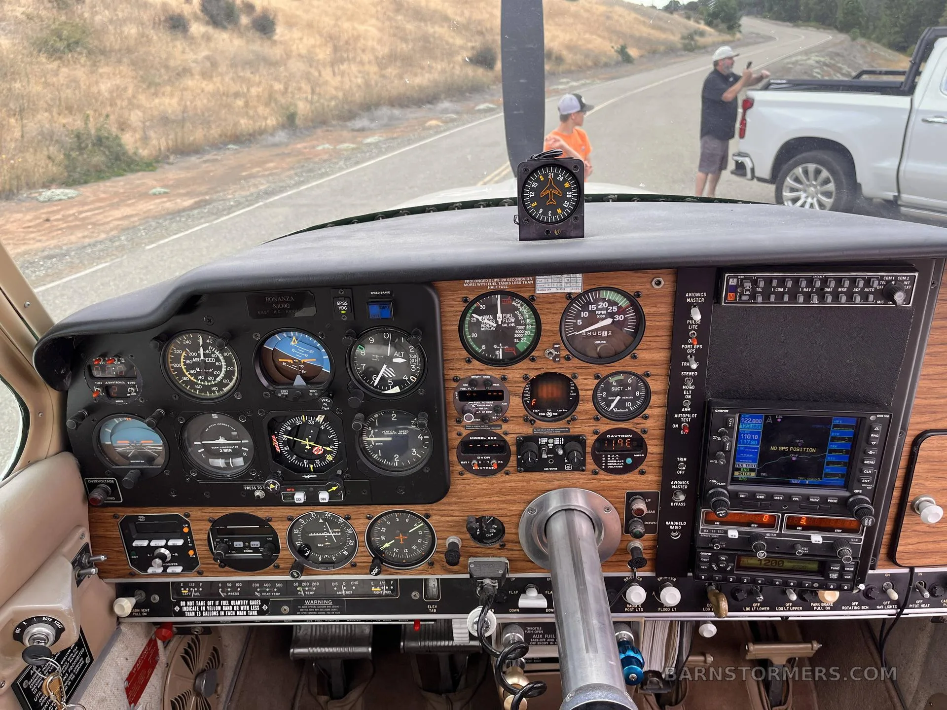 The cockpit of a Beechcraft Bonanza, showing the instrument panel with analog gauges, wooden trim, and a modern GPS unit, with people and a truck visible outside.