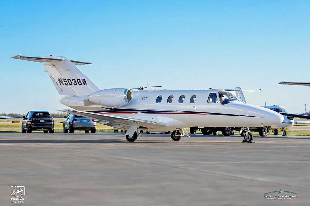 A side view of a white Cessna Citation CJ3 (N903GW) with blue and red stripes parked on an airfield with cars in the background.
