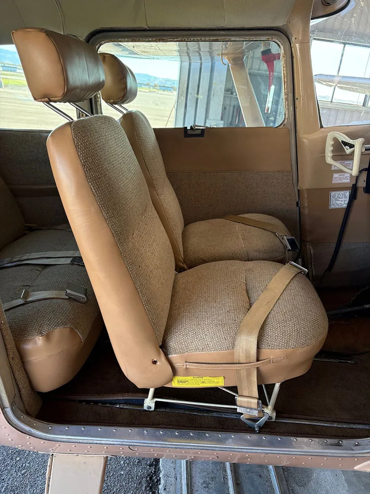 The front passenger seats of an aircraft cabin, upholstered in brown fabric and tan vinyl, with a view outside the window.