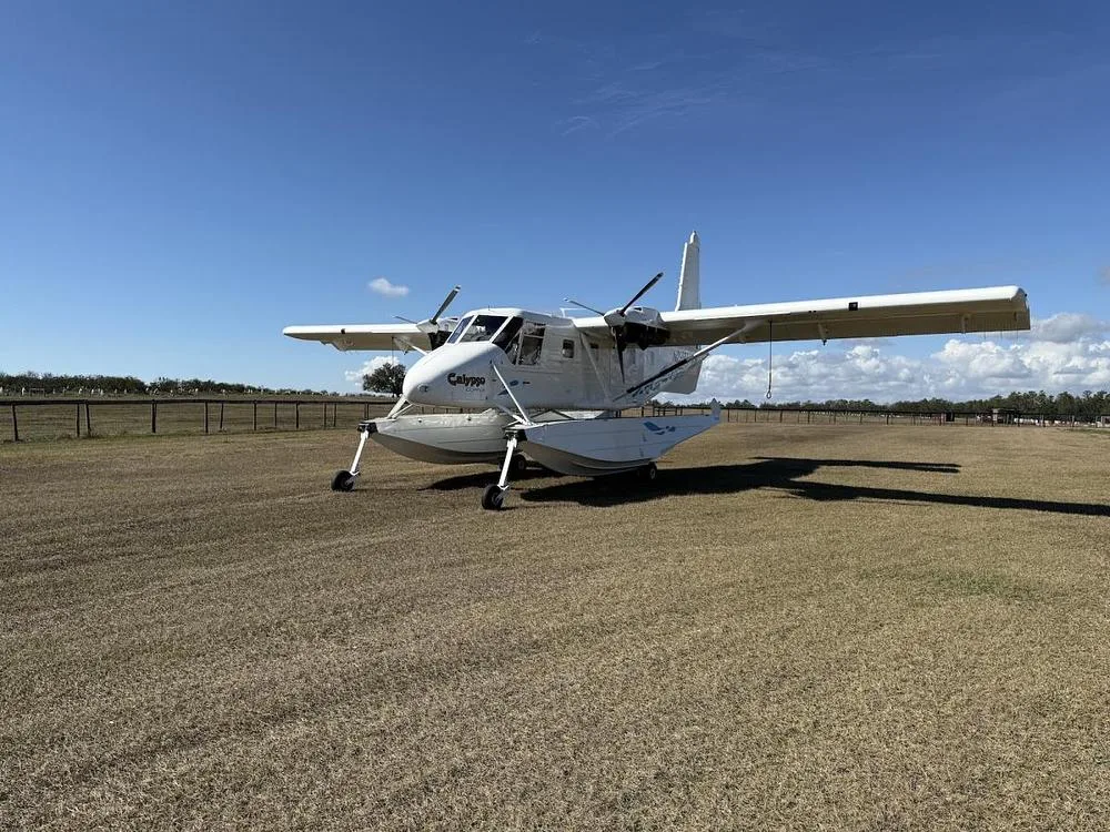 A white twin-engine seaplane with floats sits on a grassy airfield under a clear blue sky.