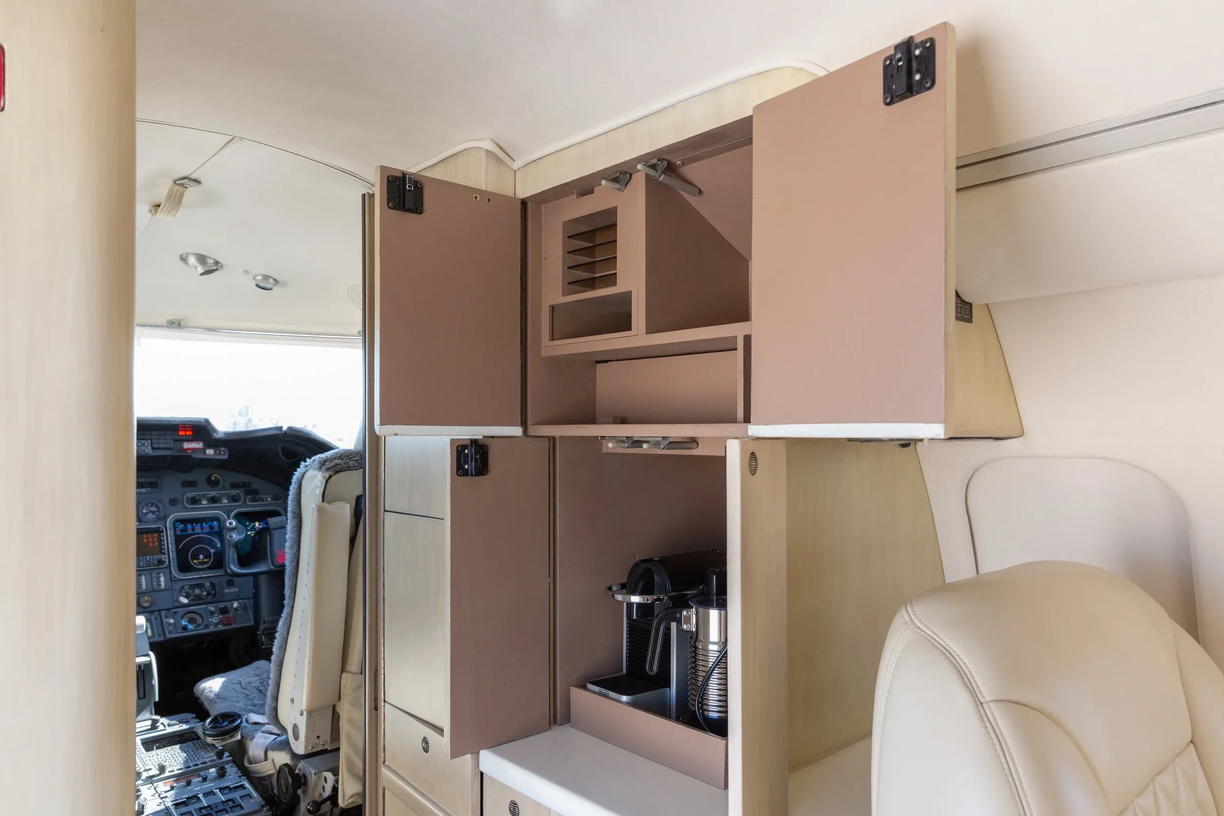 A galley area inside a private jet with open cabinets revealing a coffee machine and storage, with the cockpit visible in the background.