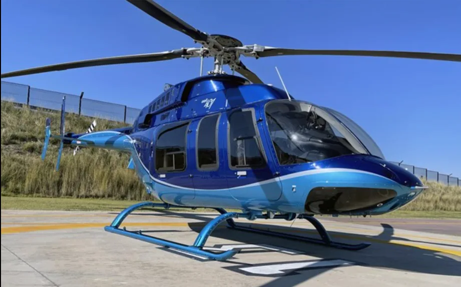 A two-tone blue Bell 407 helicopter with white accents is parked on a concrete helipad under a clear blue sky.