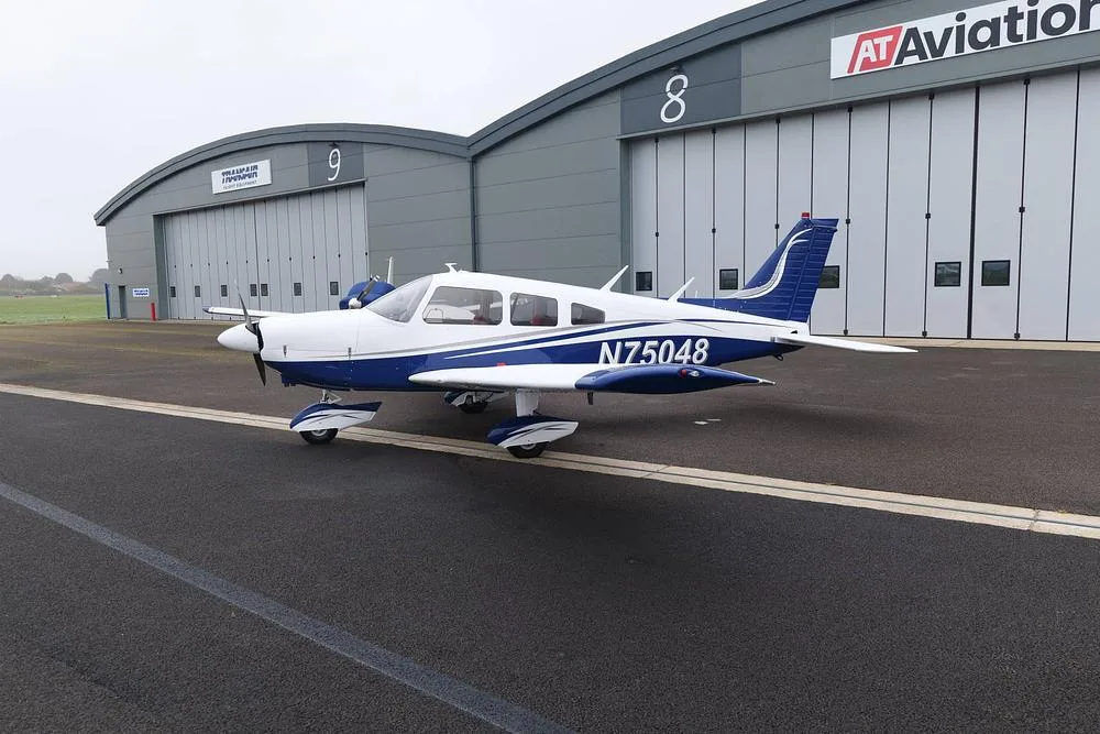 A white and blue Piper Cherokee aircraft, registration N75048, parked on an airfield in front of two hangars.
