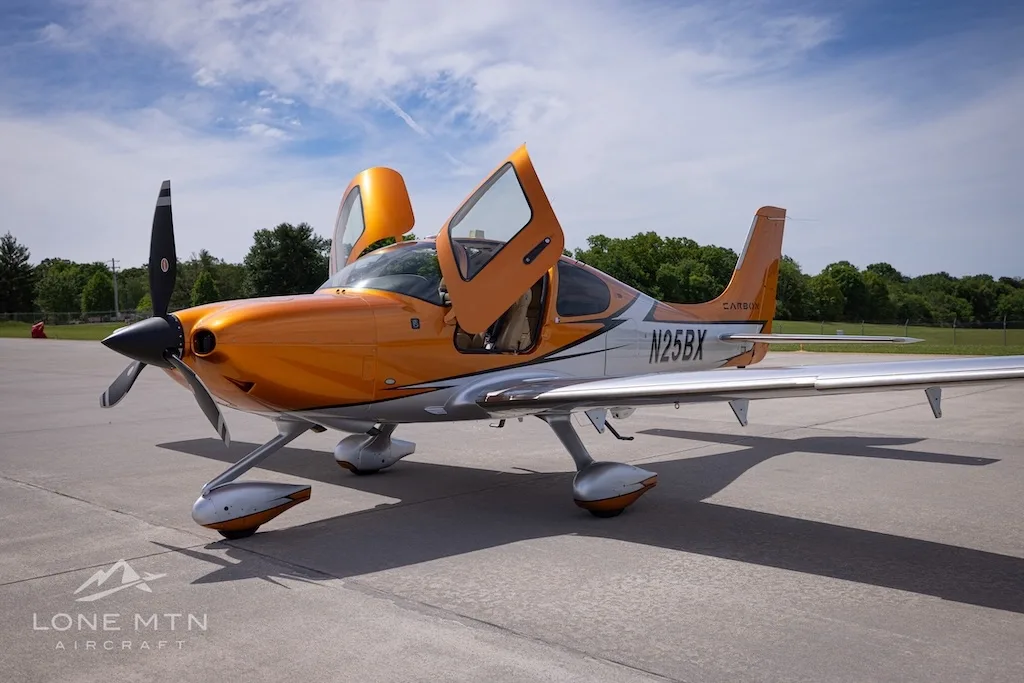 A front-left view of the orange and silver Cirrus SR22T N25BX with its doors open on an airfield.