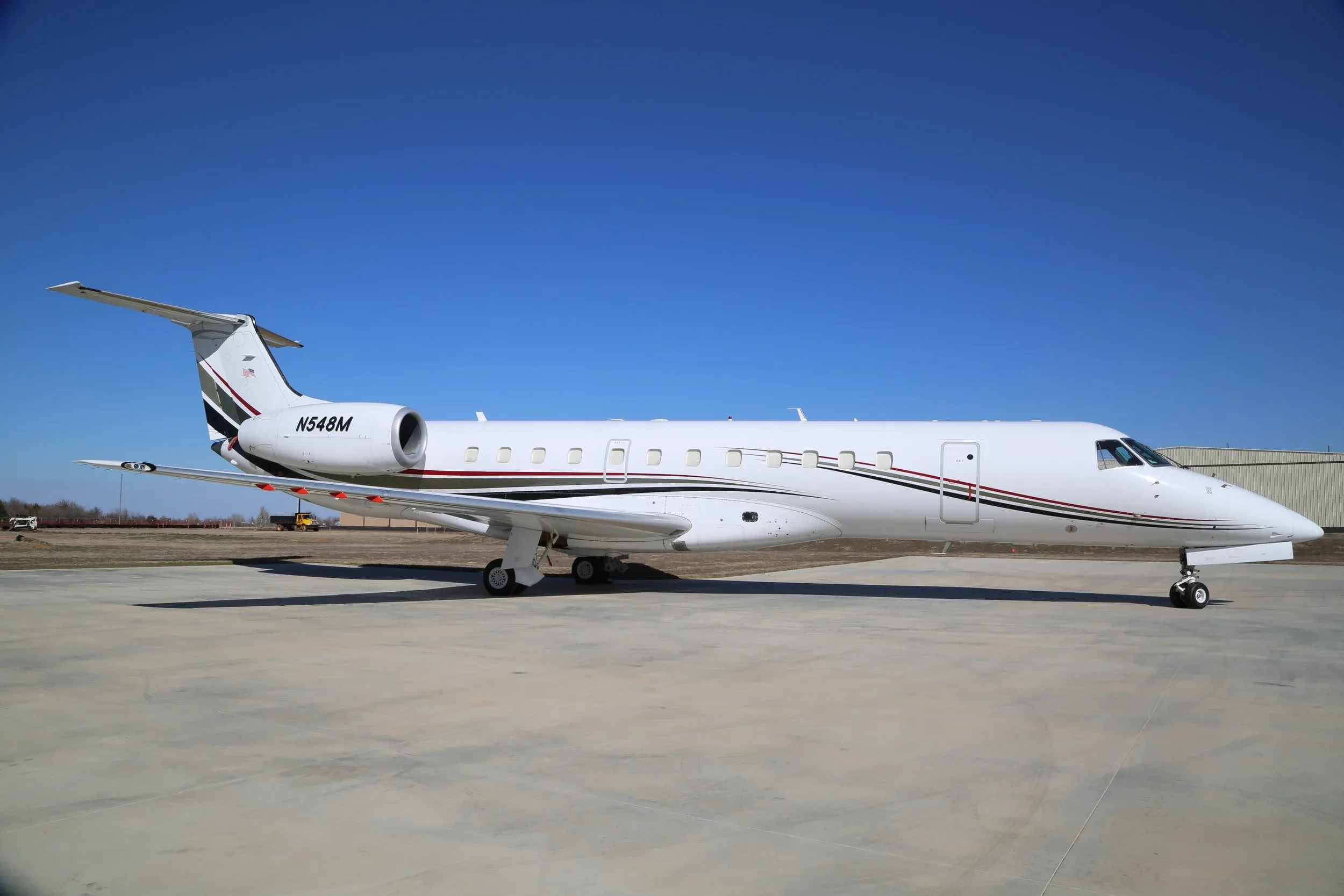 A side view of a white Embraer Legacy business jet with black, red, and gold stripes parked on a tarmac.