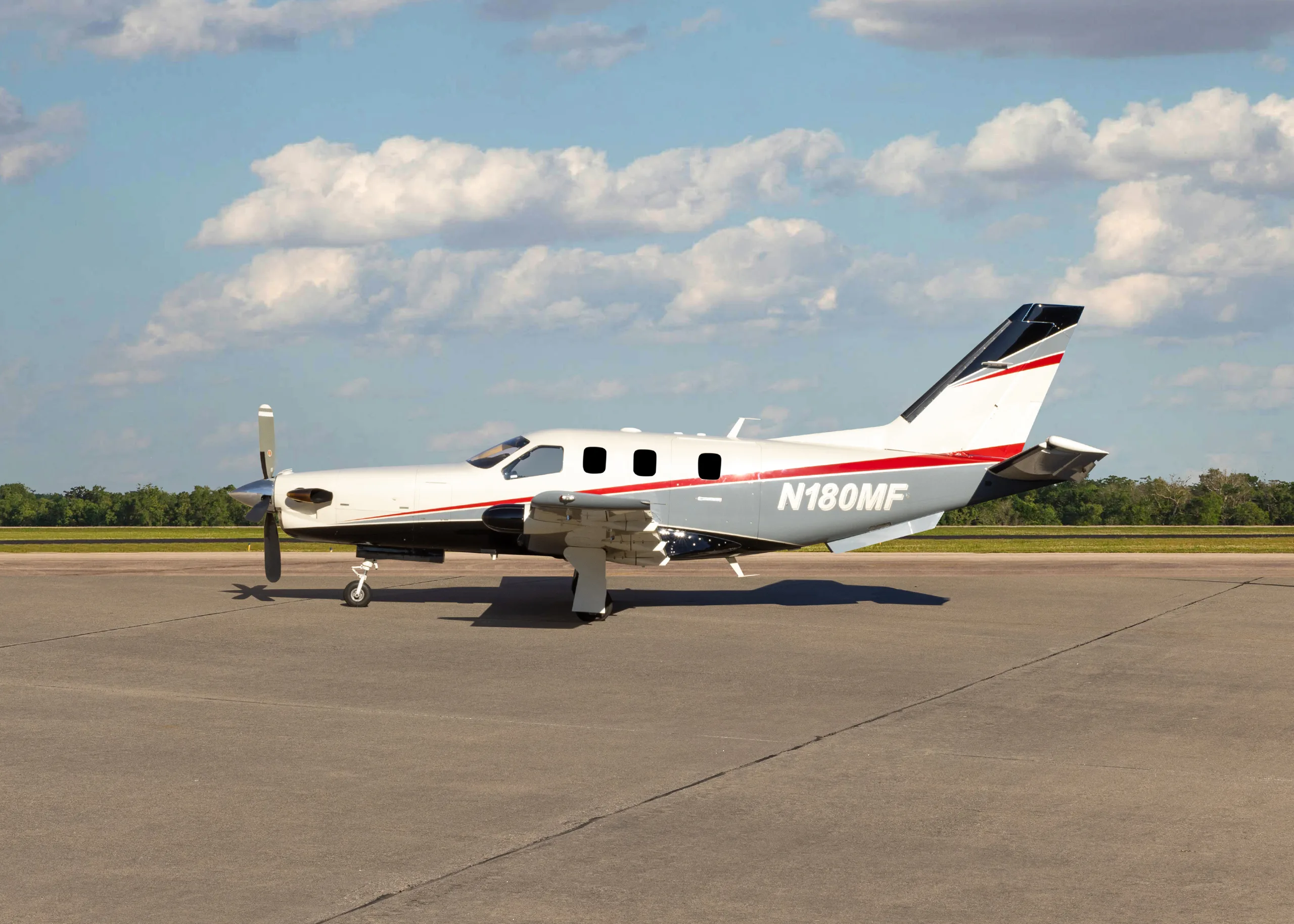 A white and grey TBM 900 series aircraft with red stripes, registration N180MF, parked on an airfield, showing the full left side.