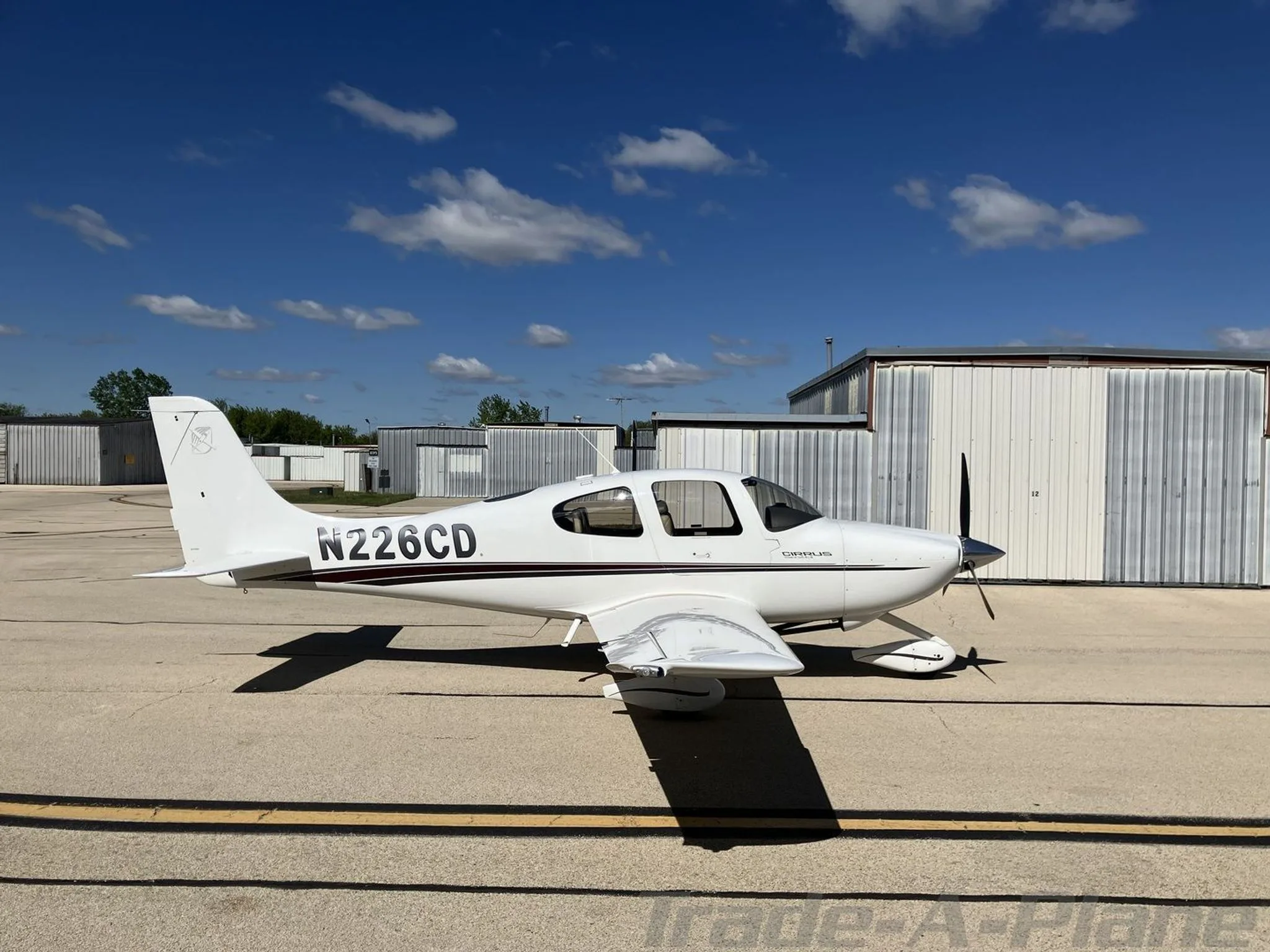 A white Cirrus SR20 aircraft with black and brown stripes, tail number N226CD, parked on an airfield tarmac with hangars in the background.